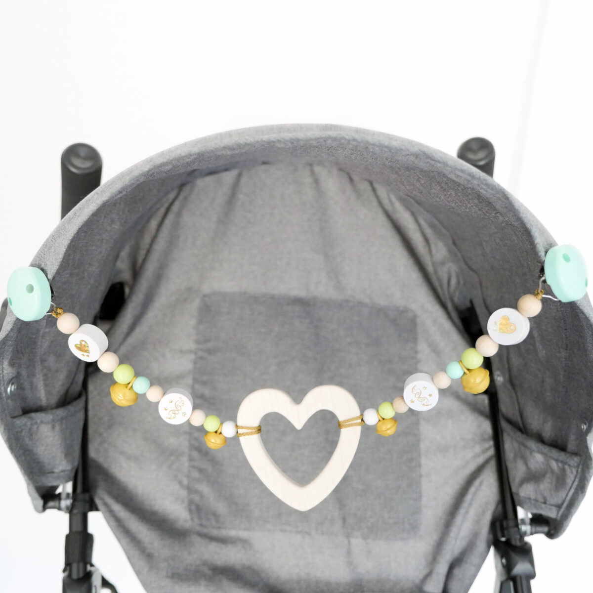 Grauer Buggy, an dessen Verdeck eine Kinderwagenkette mit Herz-Beißanhänger, Glöckchen und glitzernden Motivperlen in den Farben Natur, Mint, Weiß und Gold, hängt. Im Hintergrund ist eine weiße Betonwand.