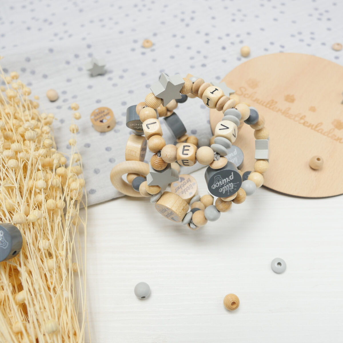 Auf einem weißen Holztisch liegen grauer, gepunkteter Stoff, beigefarbene Trockenpflanzen und ein Holzschild mit Elefanten-Logo sowie der Aufschrift "Schnullerkettenladen". Star des Fotos ist aber der mittig platzierte Greifball in den Farben Hellgrau, Grau und Natur mit dem namen "Levi", Stern-Motivperlen sowie den Motivperlen "little prince" und "little brother".