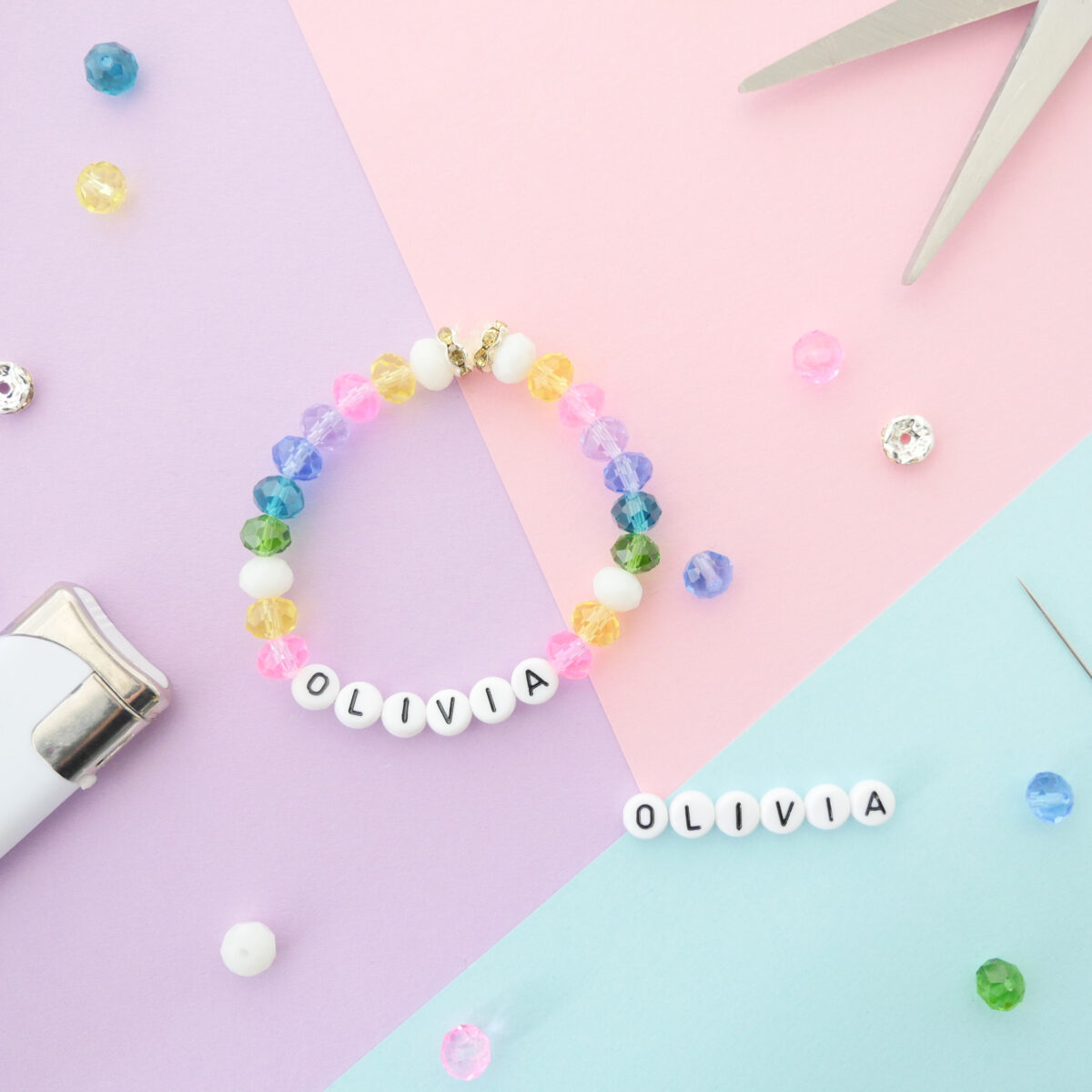 Der Untergrund besteht aus drei bunten Blättern Papier in Flieder, Rosa und Hellblau. Darauf liegt ein buntes Kinderarmband aus Acrylglasperlen mit dem Namen "Olivia". Drumherum liegen weitere Perlen, ein Feuerzeug und eine Schere.